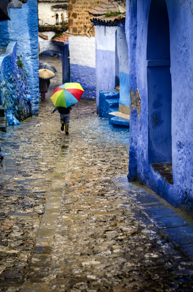 Niño corriendo con paraguas de colores entre las calles azules de ChefChaouen, Marruecos. Fotografía de Milena Martínez, fotógrafa en Madrid